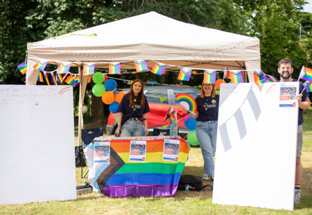 Three members of Futures staff stood around a Pride stall in a Gazebo, flanked with canvases and covered in brightly coloured rainbow flags.